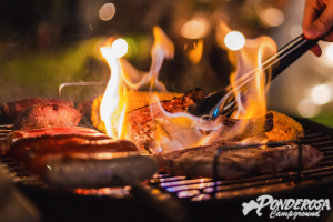Steak grilling over open flames on a barbecue, with tongs and bokeh lights in the background.