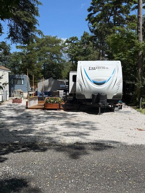 Camper trailer parked in a tree-shaded gravel lot beside planters and a small building