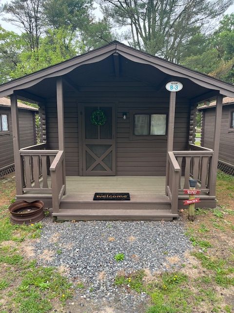 Small brown cabin porch with steps, railing, wreath on door, and gravel path in a wooded setting