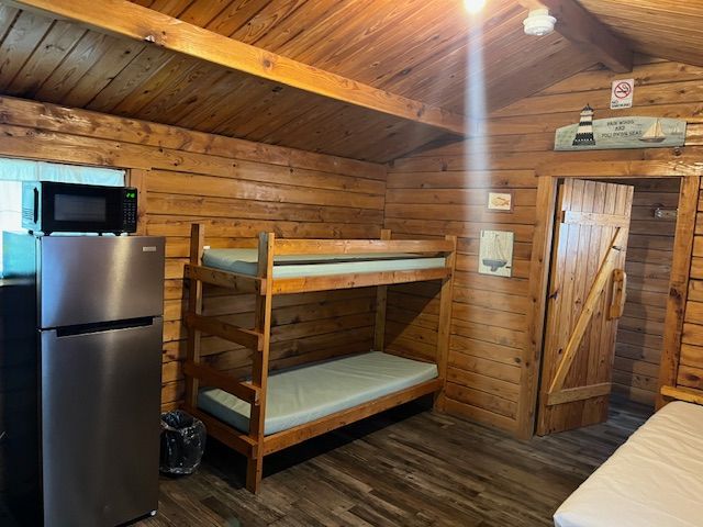 Rustic cabin bedroom with bunk bed, refrigerator, microwave, wooden walls, and loft-style ceiling