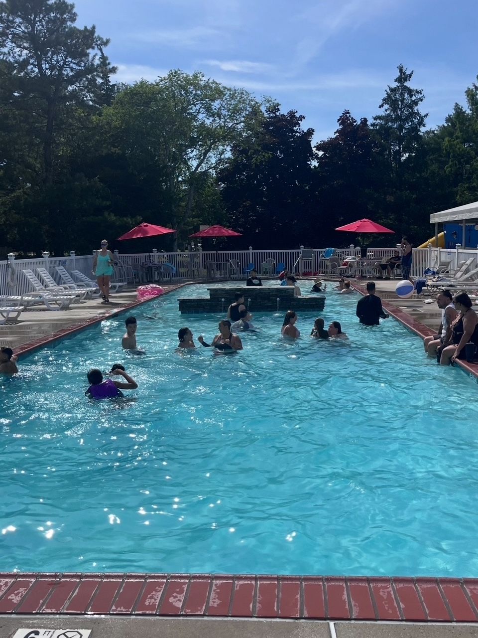 Crowded outdoor swimming pool with people swimming and lounging under red umbrellas on a sunny day