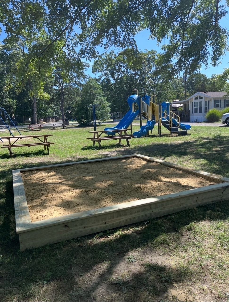 Playground with blue slides, picnic tables, and a sandy sandbox under trees beside a building.