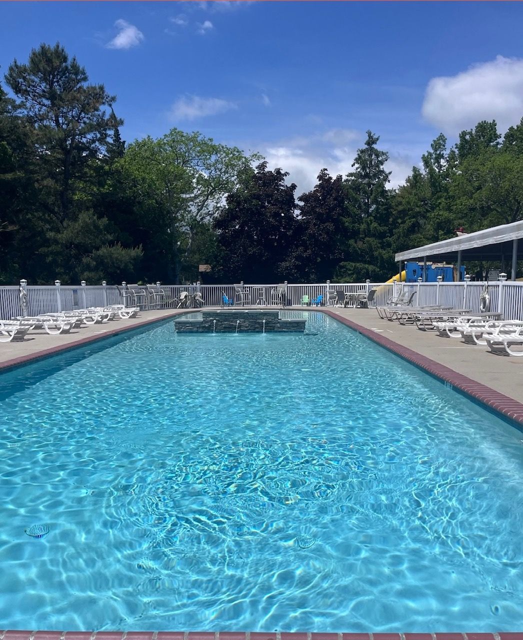 Outdoor swimming pool with clear blue water, white lounge chairs, and trees under a sunny sky