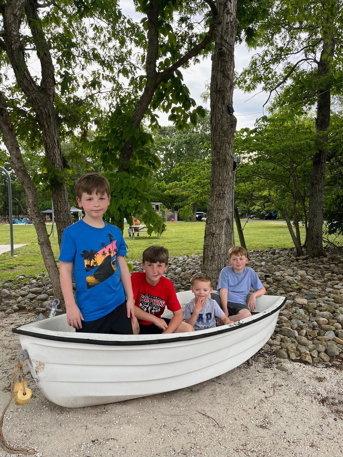 Three children sitting in a white boat-shaped planter under trees in a gravel park.