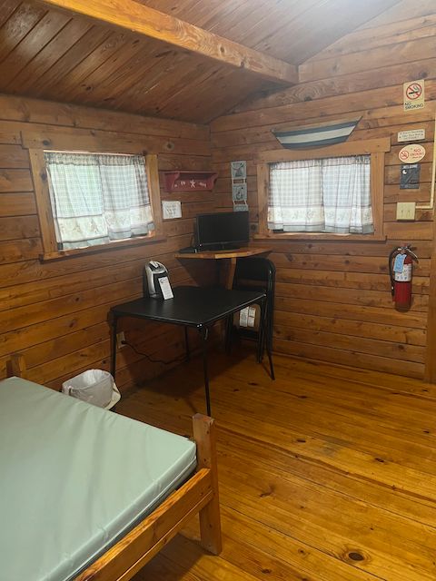 Rustic wood-paneled bedroom with bed, desk, TV, window, and wall-mounted fire extinguisher
