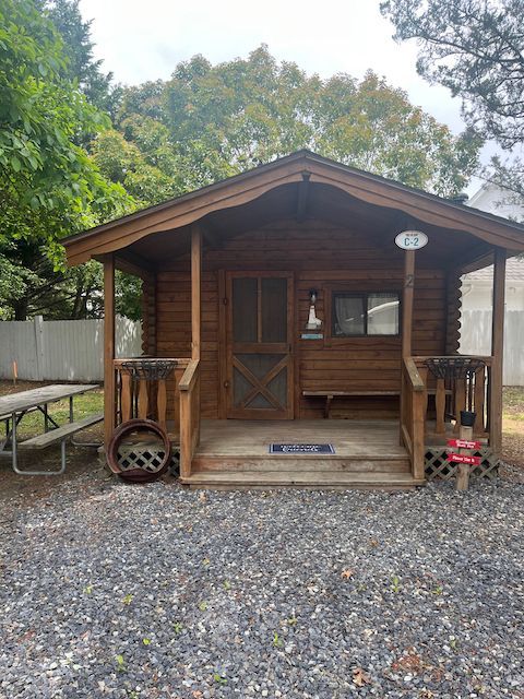 Small rustic wooden cabin with porch, gravel foreground, and trees in the background