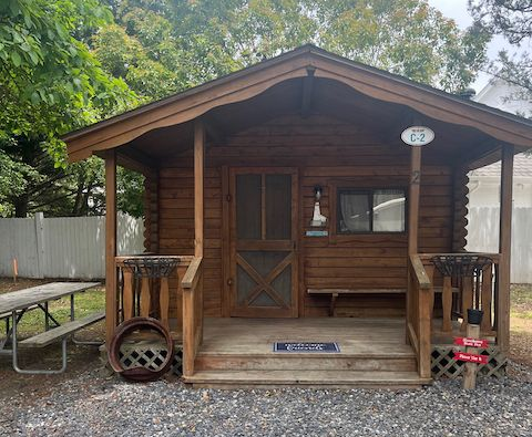 Small brown cabin with a front porch, centered front door, and gravel path in a wooded setting