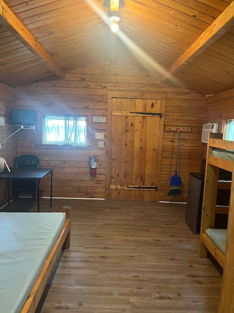 Wood-paneled cabin bedroom with bunk bed, window, and wooden door under a sloped ceiling.