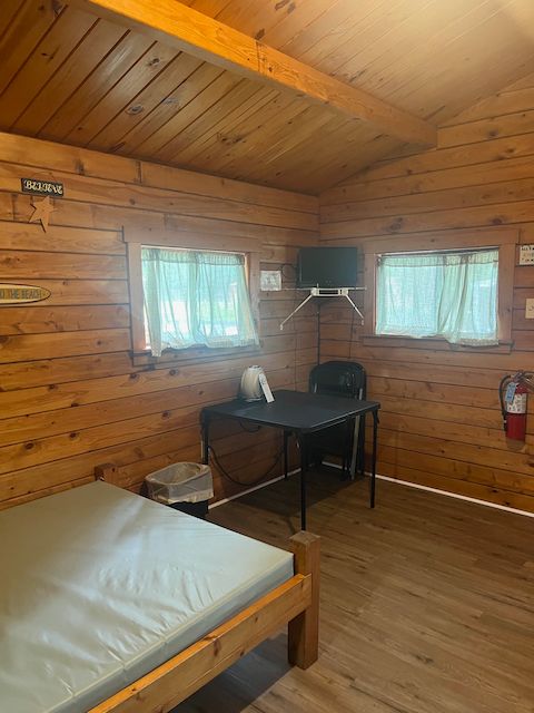 Wooden cabin bedroom with bed, desk, chair, and two windows with curtains