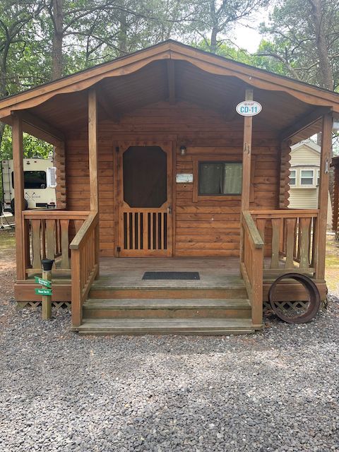 Small brown cabin with a front porch, centered front door, and gravel path in a wooded setting