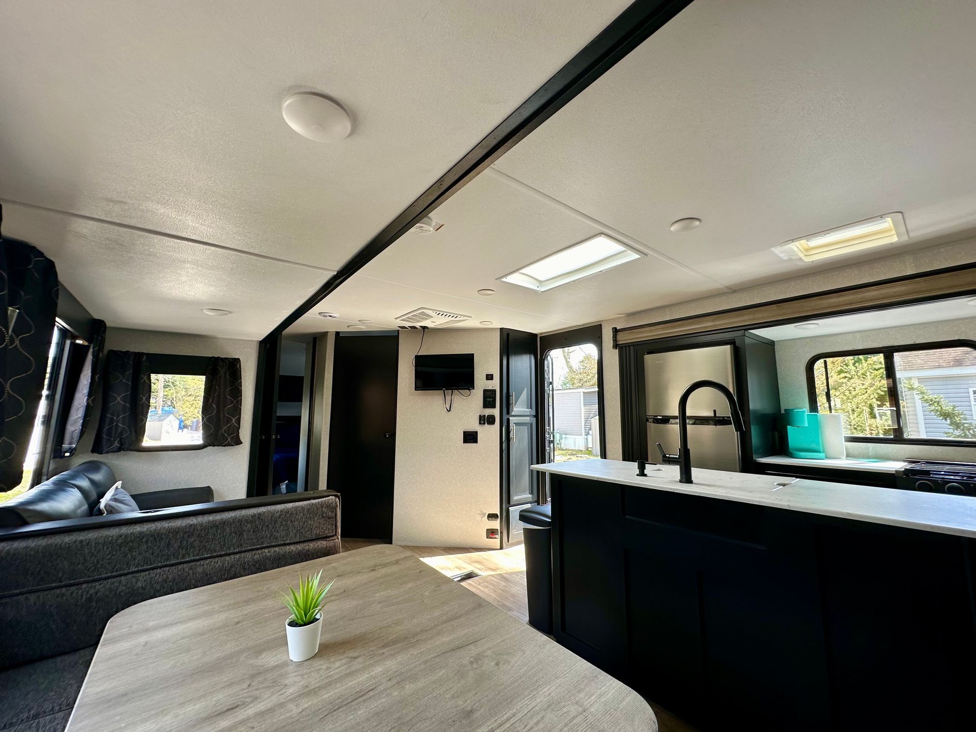Modern RV interior with black kitchen, gray sofa, and large windows showing greenery