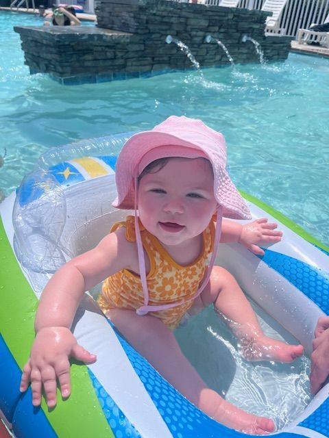 Child in pink sunhat sitting in a colorful inflatable pool float in a swimming pool, smiling in the water.