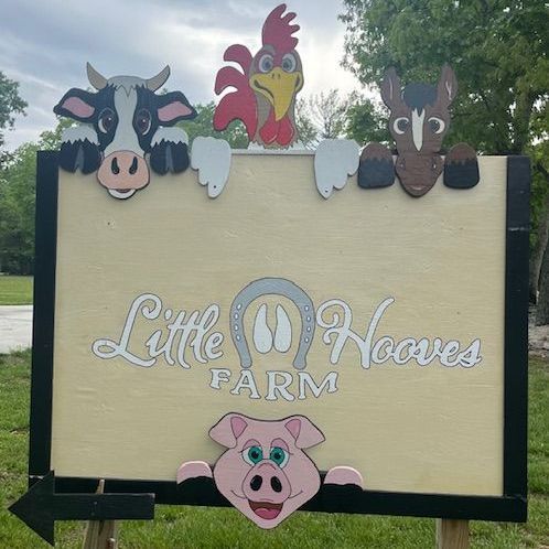 Little Hooves Farm sign with cartoon cow, chicken, goat and pig against a wooded outdoor background