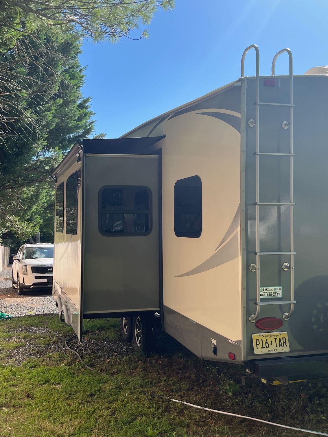White RV trailer parked on grass beside trees, with an open side slide-out and rear ladder.