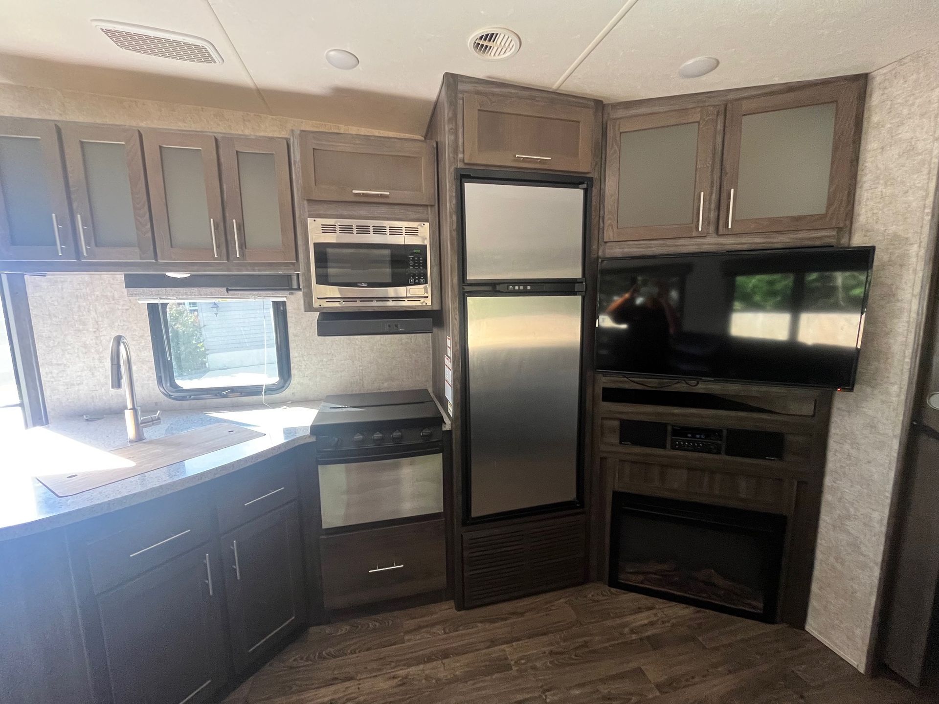 Compact RV kitchen with dark cabinets, stainless appliances, sink, and built-in TV and fireplace