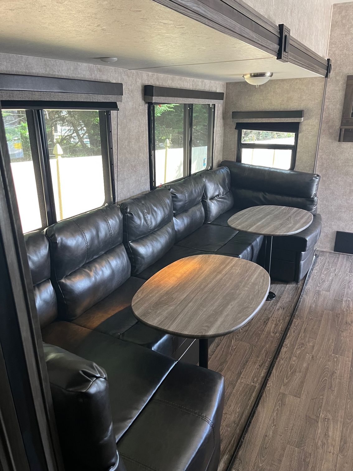 Interior of an RV with black leather bench seating and round wood tables beside large windows.