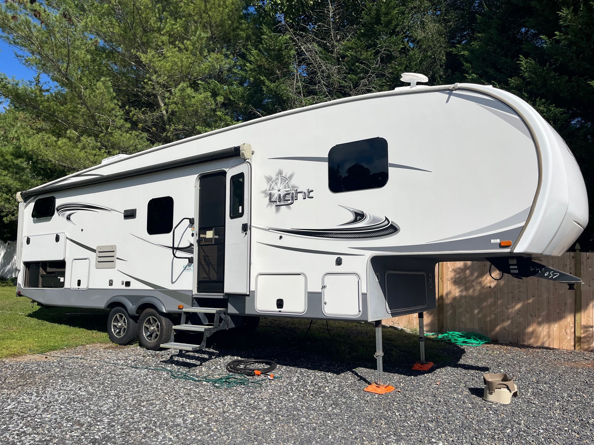 White fifth-wheel RV parked on gravel beside a wooden fence and trees, with the door open.