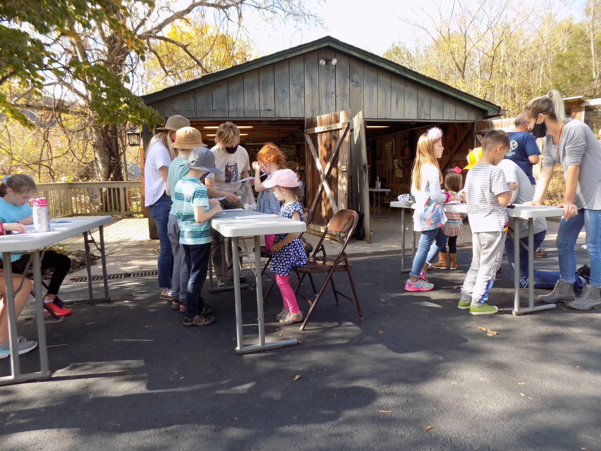 Children and adults at outdoor tables, engaged in an activity in front of a weathered wooden building.