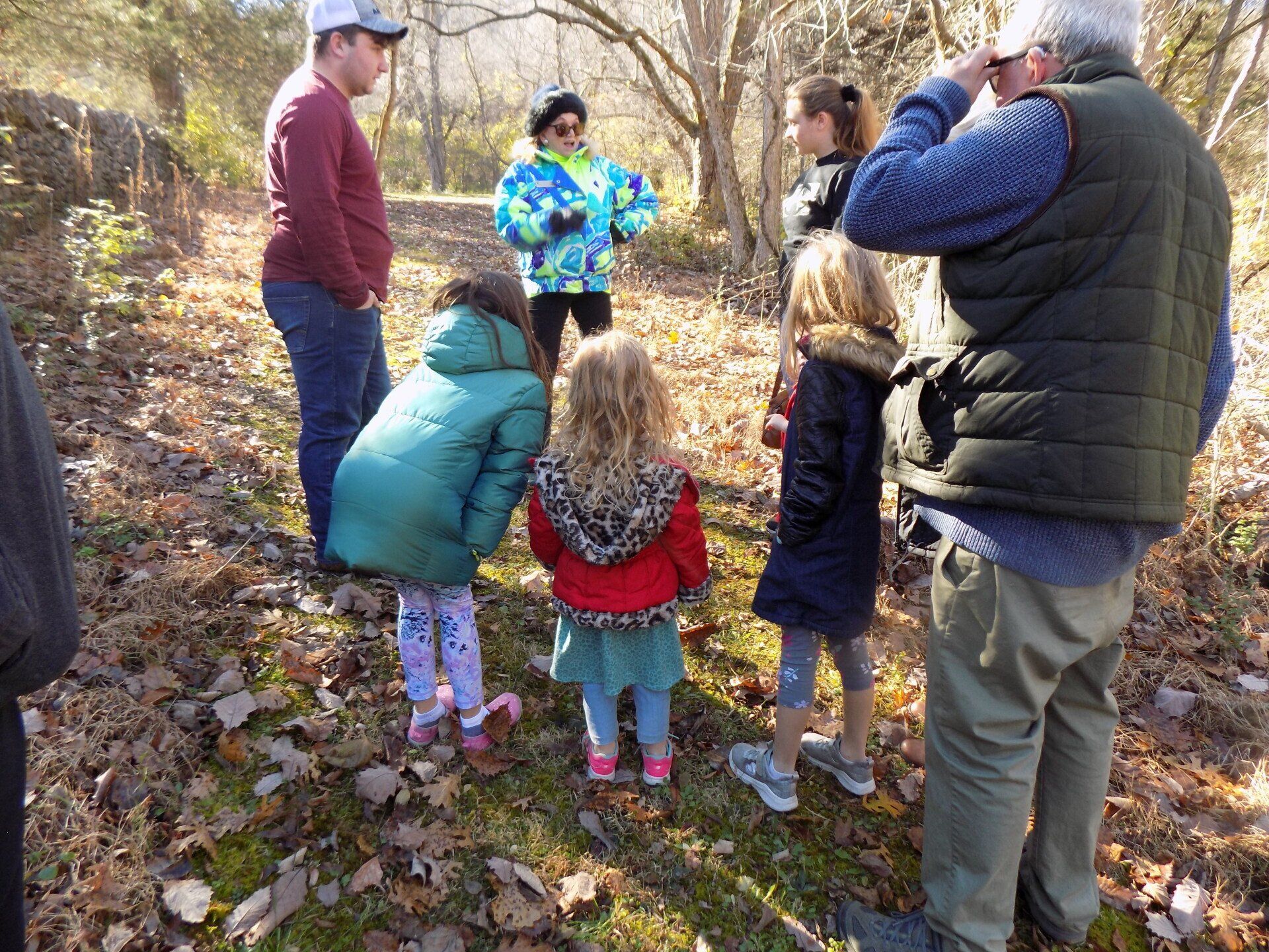 Group of people outdoors in a wooded area; some children in colorful coats, looking at something on the ground.