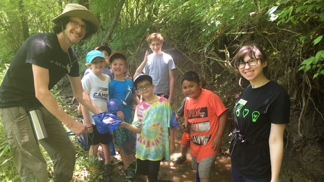Group of children and adults exploring a stream. Some hold nets, smiling in a wooded area.