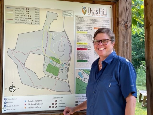 Woman smiles next to a map of Owl's Hill Nature Sanctuary. The map is outdoors in a wooden kiosk.