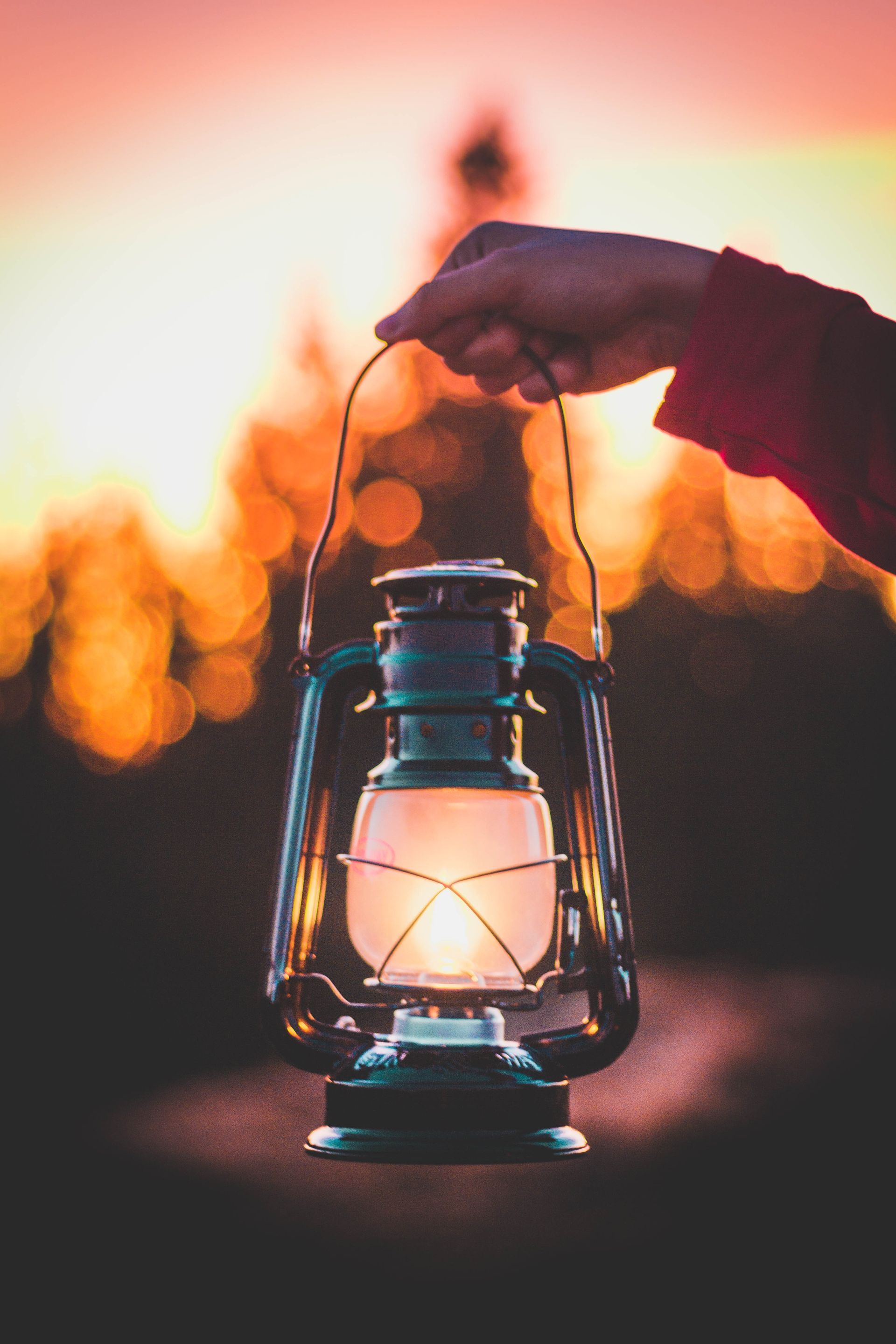 Hand holding a lit lantern against a blurred sunset backdrop.