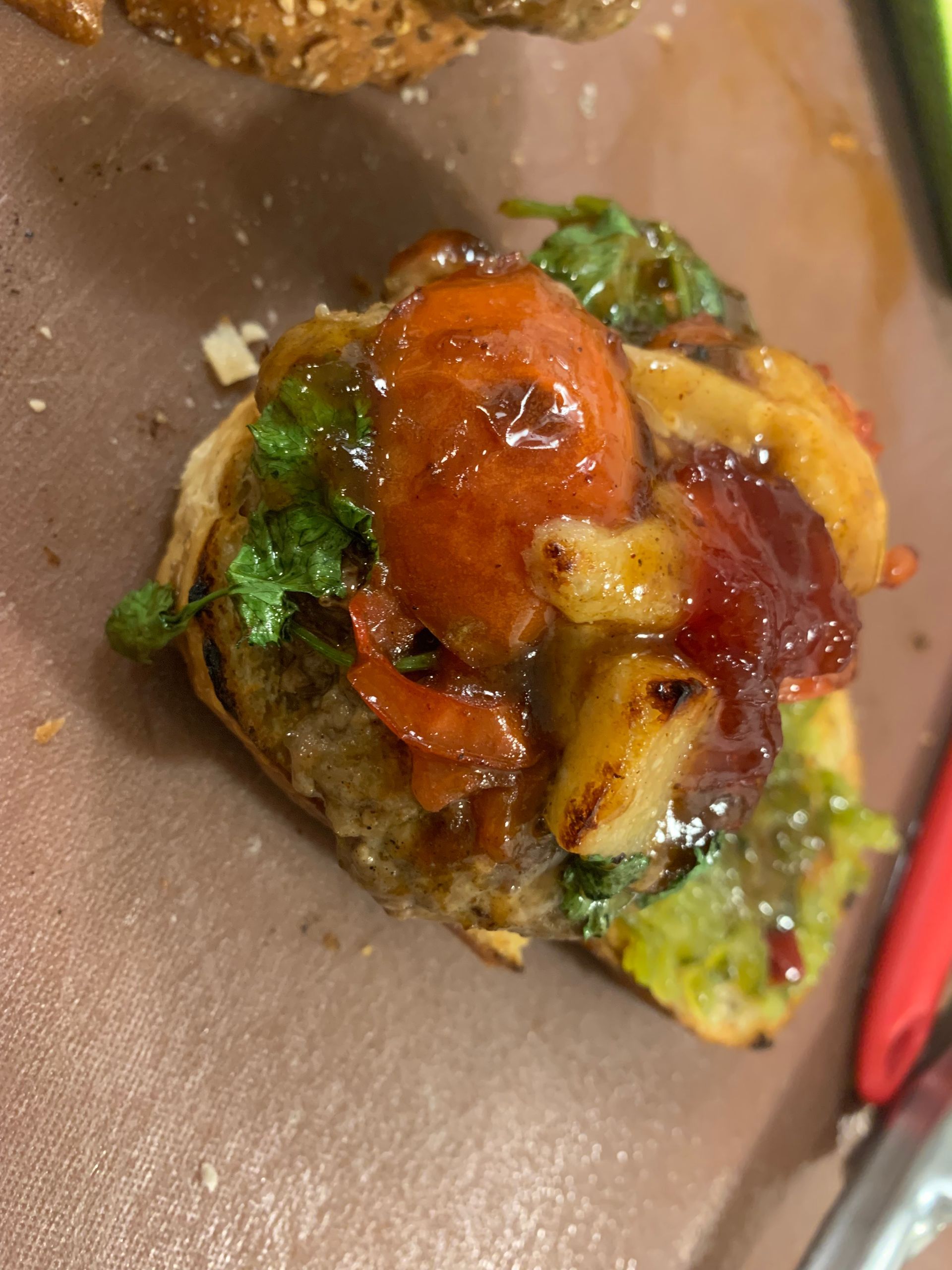 Close-up of a messy burger patty topped with sauce, melted cheese, and greens on brown paper.