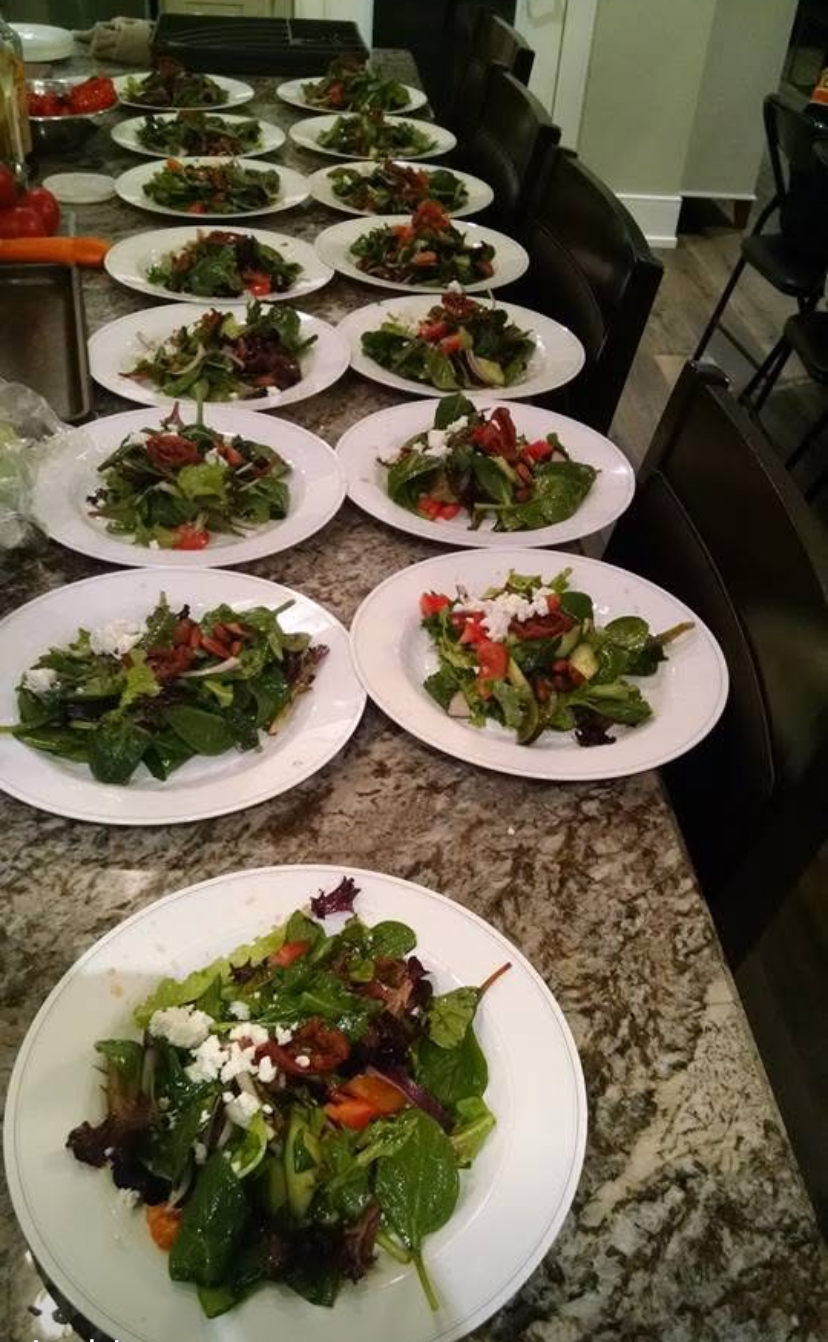 Plated salads lined up on a kitchen counter, with mixed greens and tomatoes on white plates.