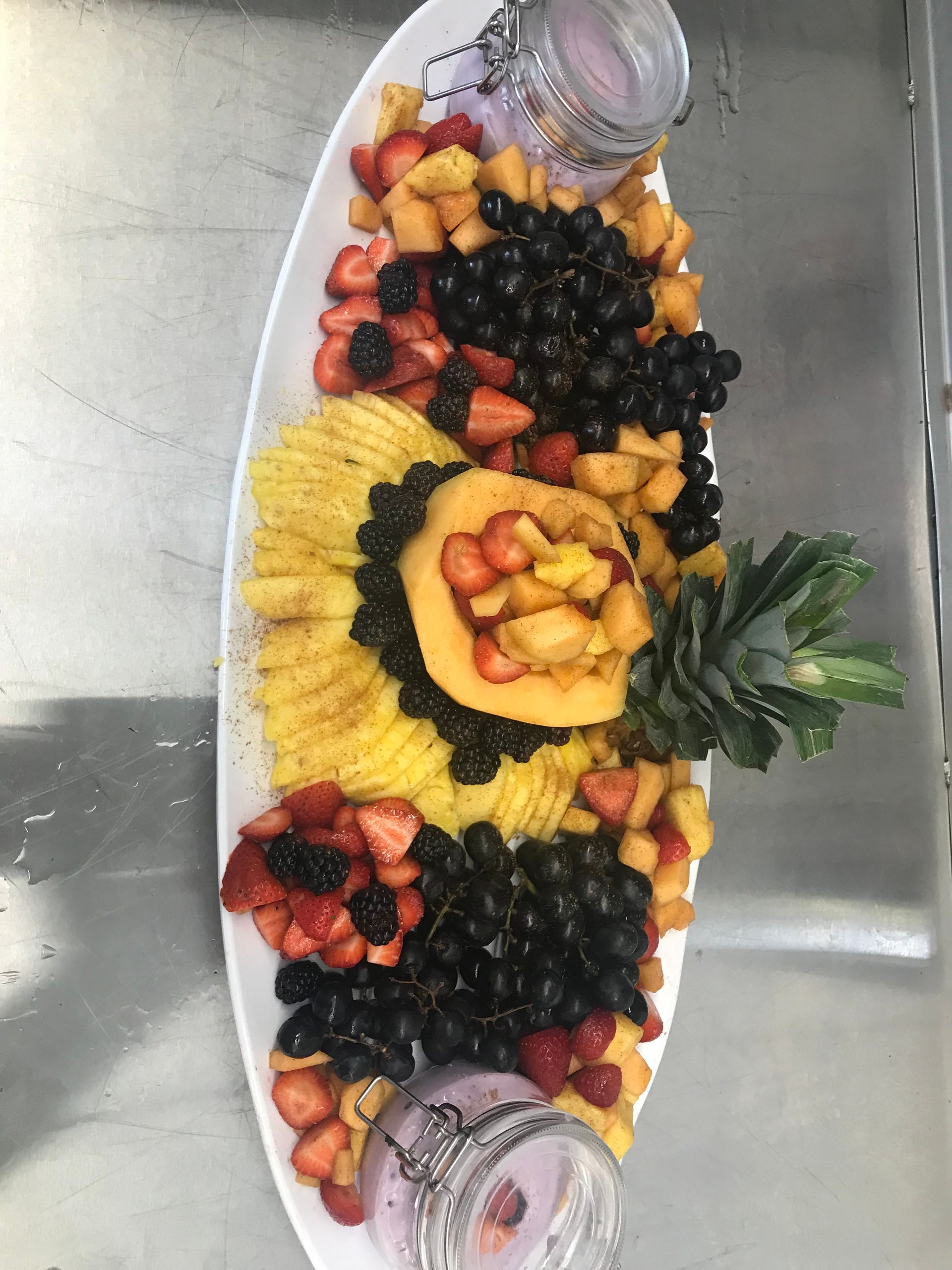 Fruit platter arranged in a pineapple shape on a tray, with grapes, berries, pineapple, and two mason jars