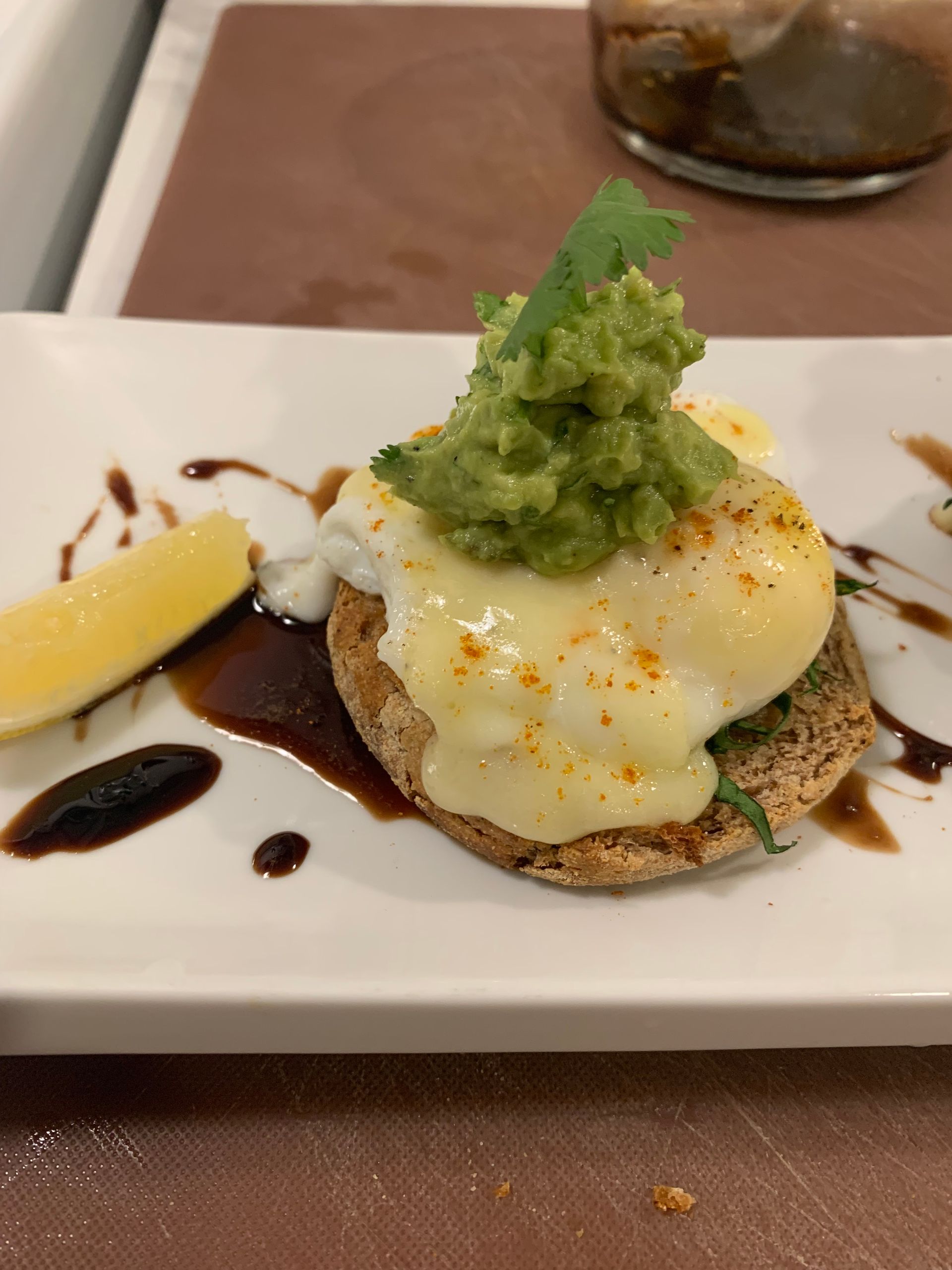 Eggs Benedict with avocado garnish on a white plate, drizzled with sauce and served with a lemon wedge.