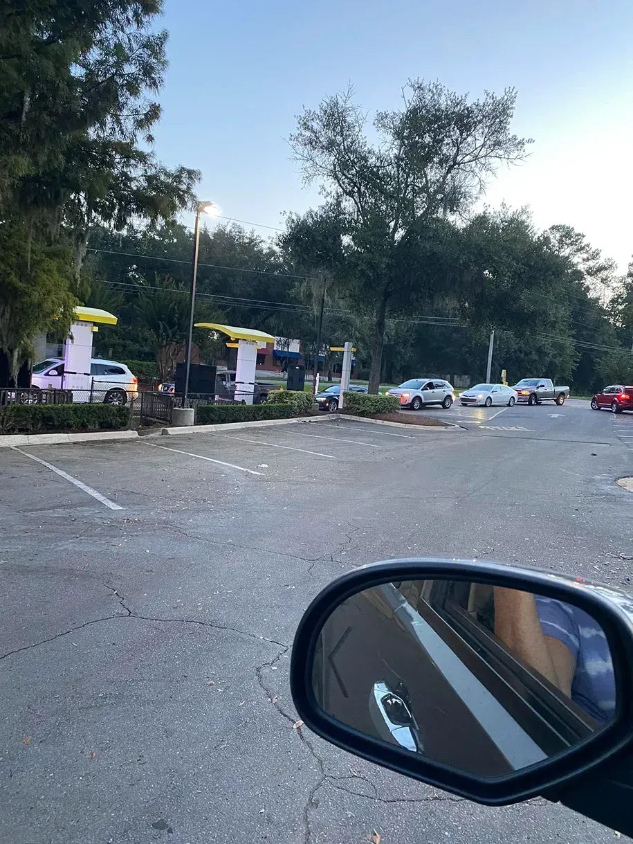 Parking lot with scattered litter, several cars, and trees at dusk, viewed from a car side mirror.