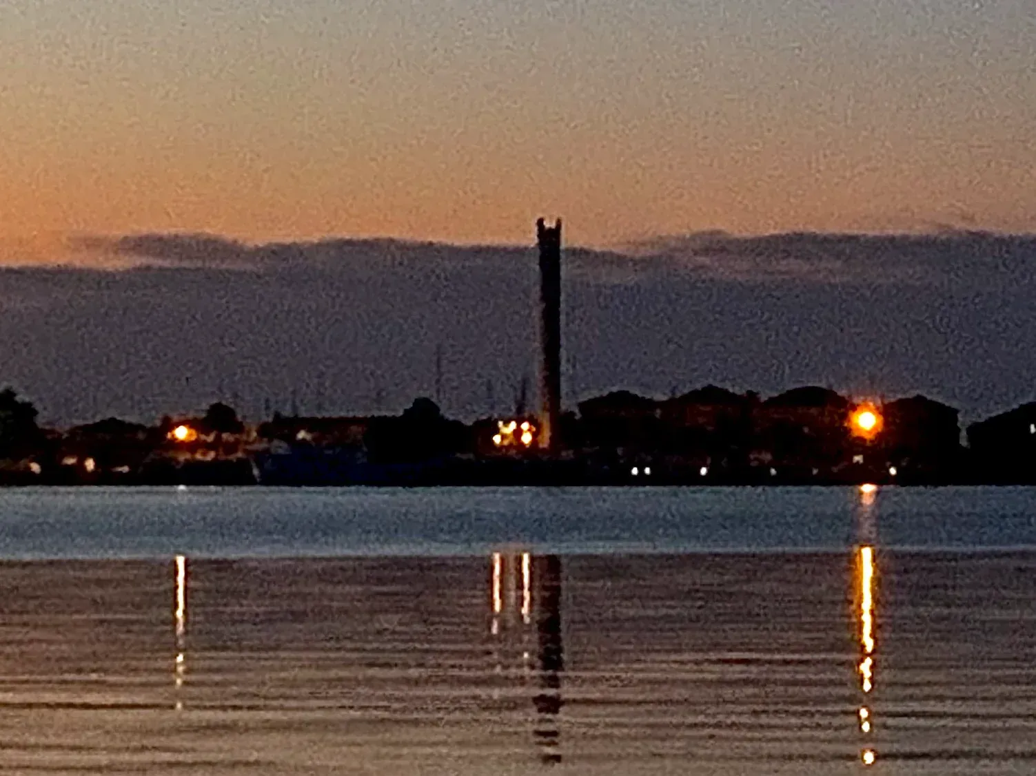 Dusk over a calm waterfront with silhouetted buildings and a tall tower reflecting on the water