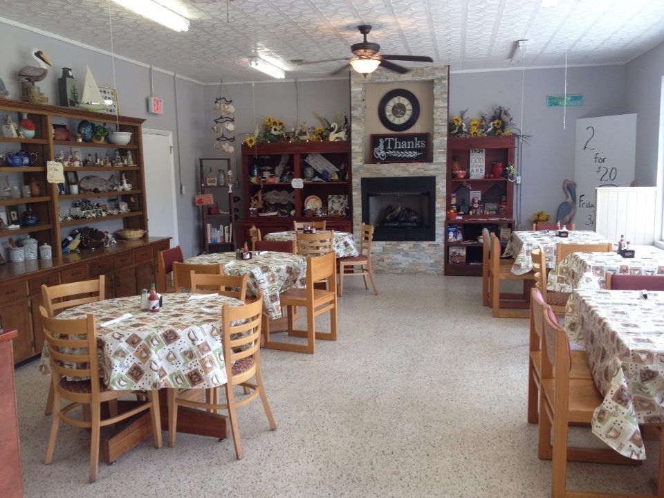 Small cozy dining room with wooden tables, patterned tablecloths, and shelves of decor around a fireplace.