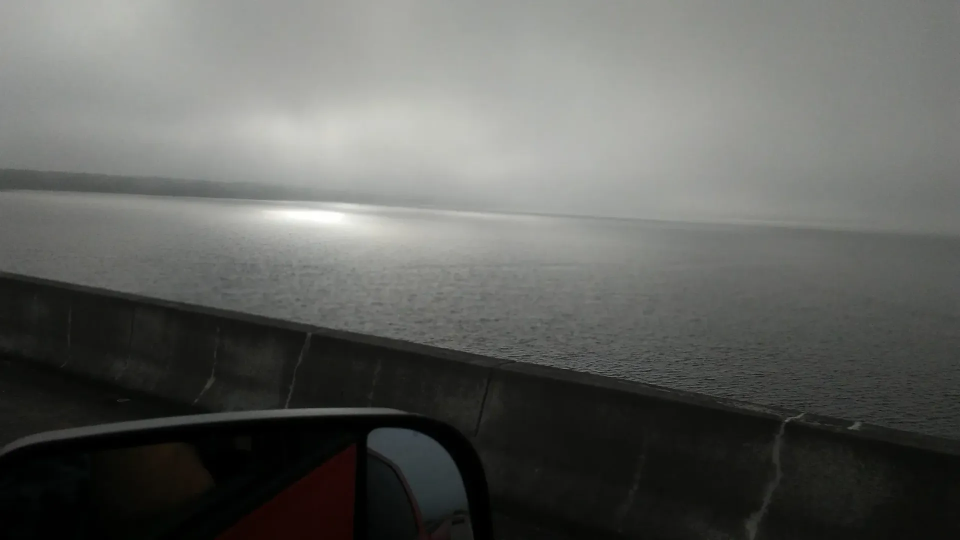 Rainy highway seen from a car, with a wet guardrail and gray sky over water.