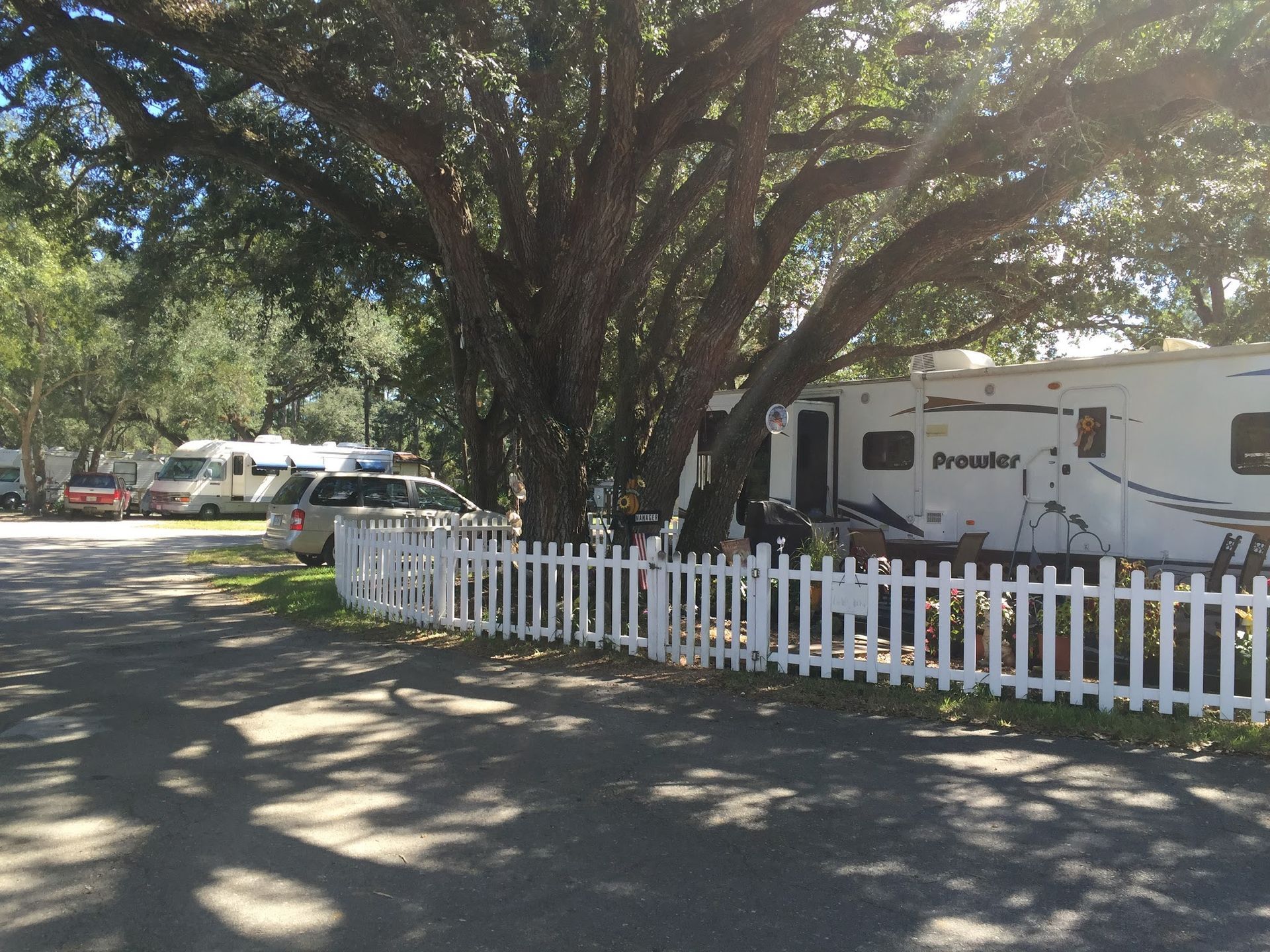 White RV parked under large trees behind a white picket fence in a shaded campground.