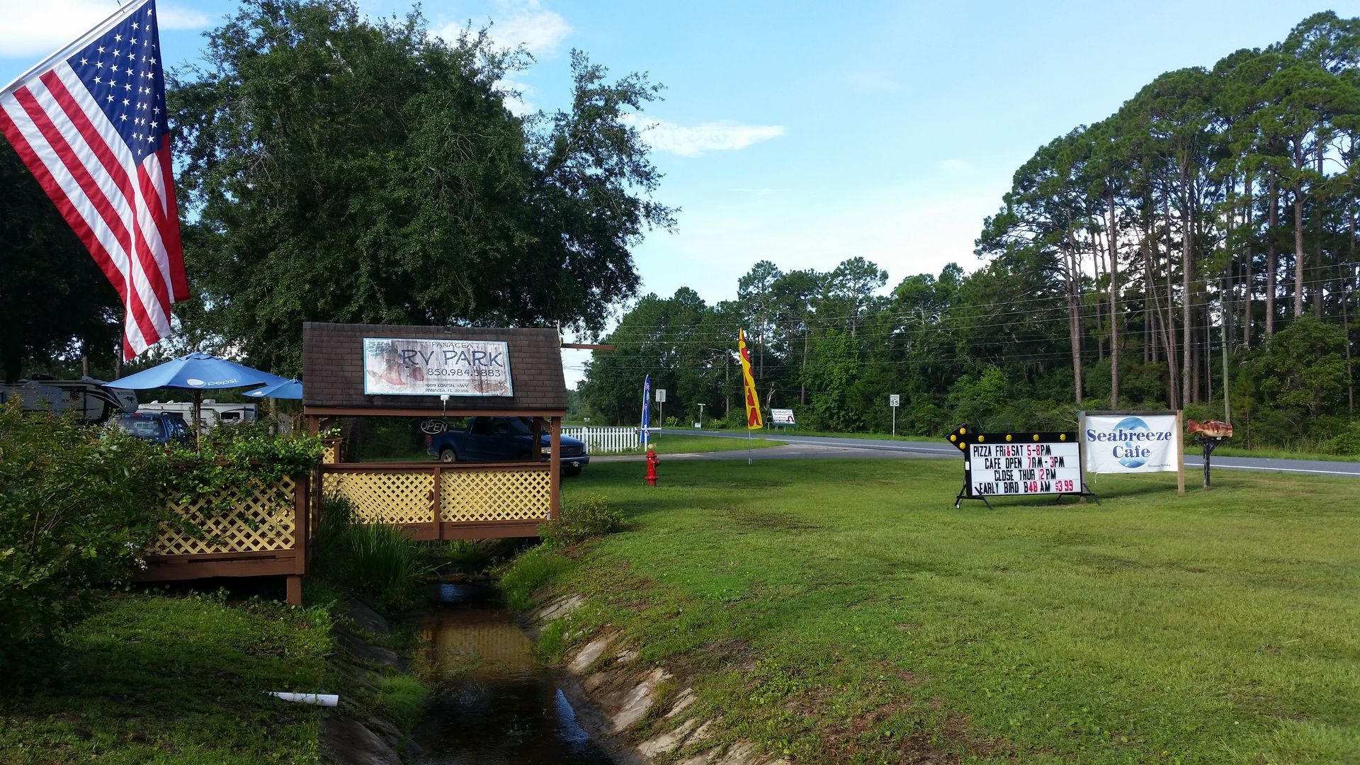 American flag and park entrance sign beside a grassy field and small creek under a sunny sky