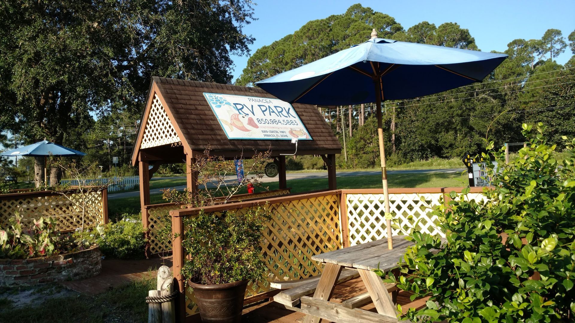 Small garden café with a blue umbrella, wooden sign, and fenced seating area in sunlight