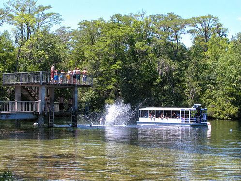 Tour boat on a river near a viewing deck as a fountain splashes beside the shoreline