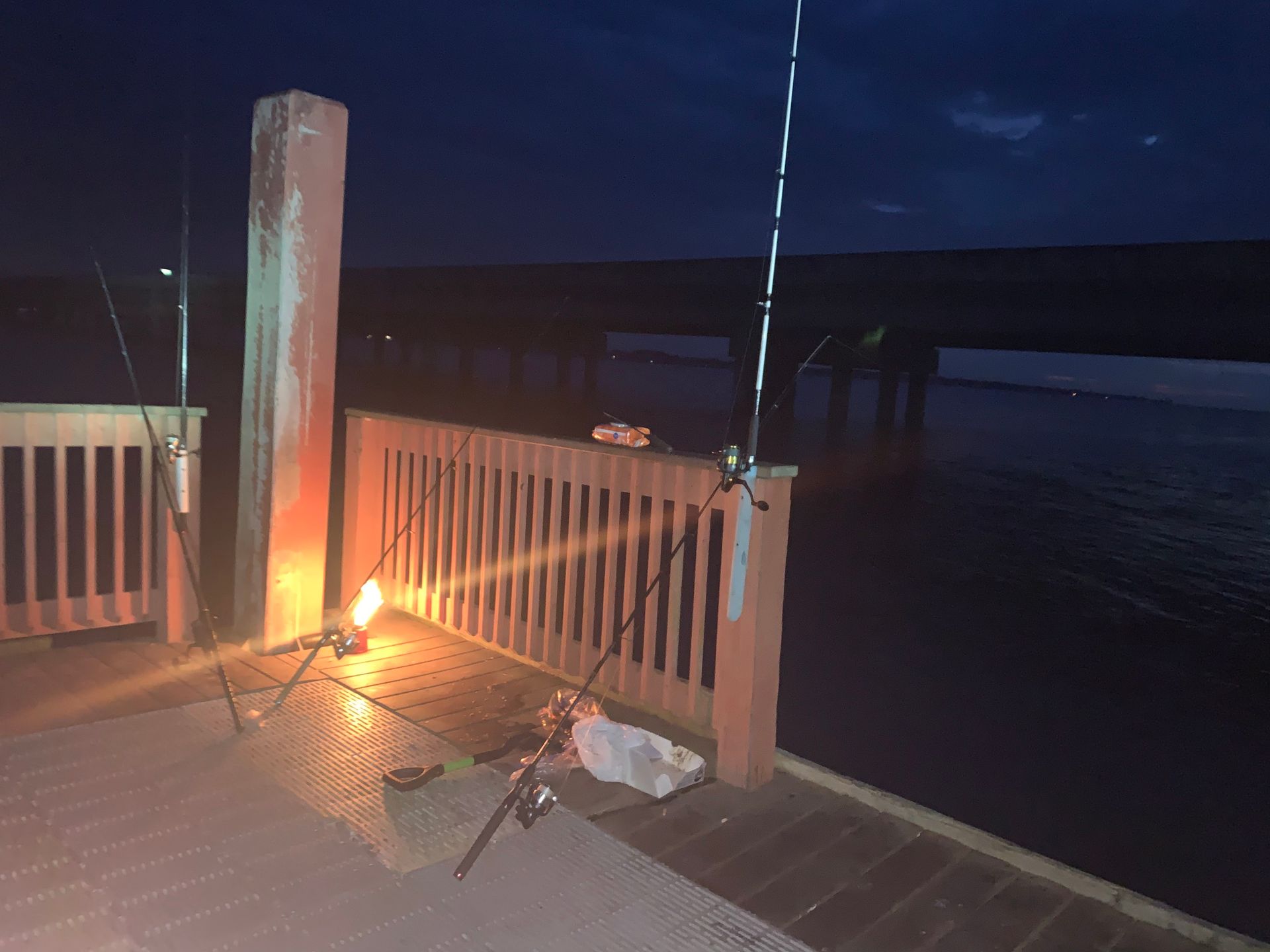 Night dock with a lit torch beside a white gate, fishing rod, and calm water beyond.