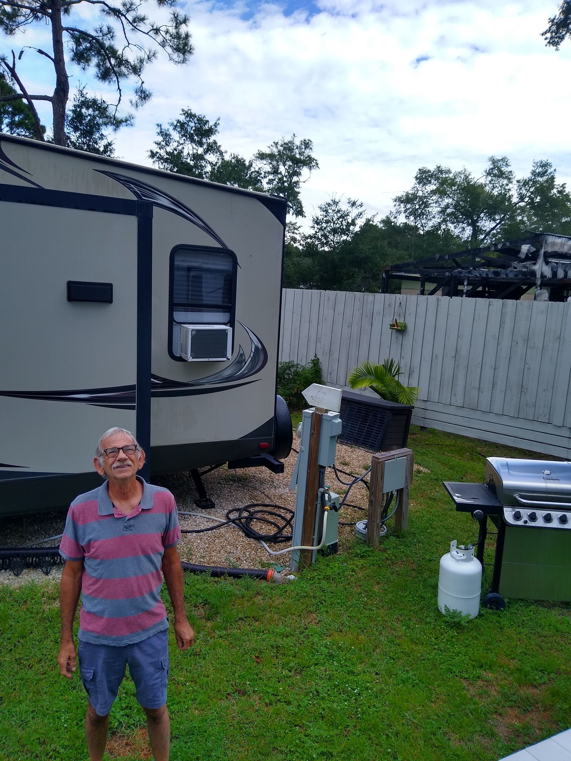 Person standing beside a white camper in a grassy yard with a fence, grill, and propane tank.