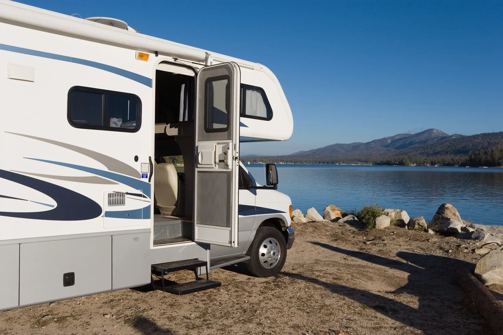 White RV parked by a lakeshore with its door open, mountains in the background.