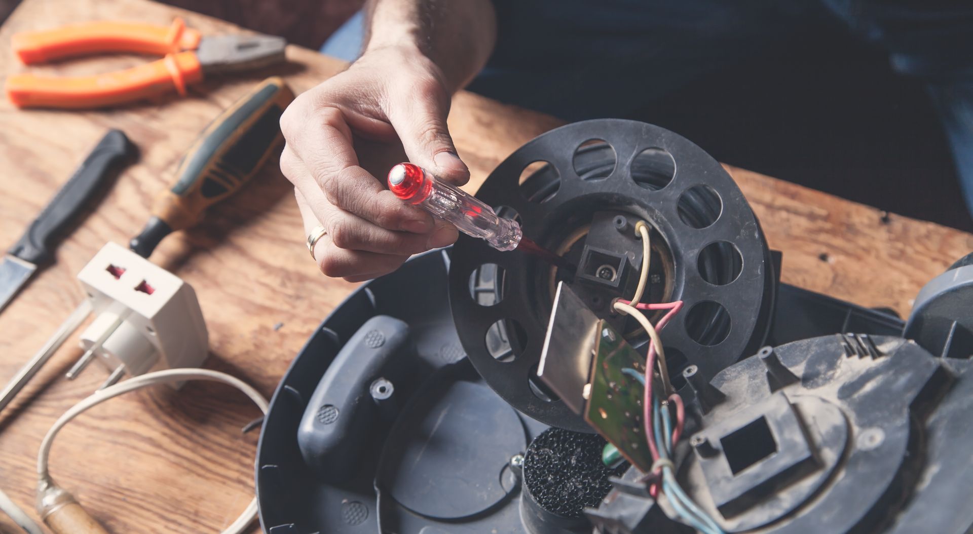 Hands repairing a black electric reel with red wires on a wooden workbench, tools nearby