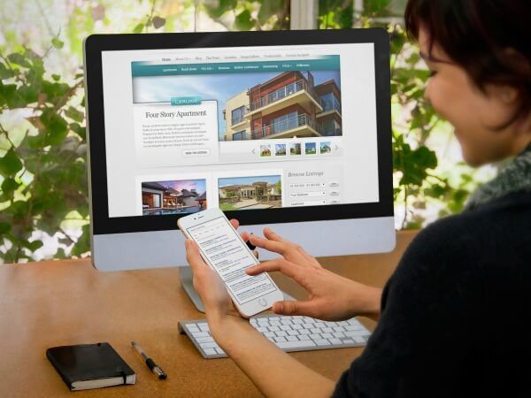 A woman is looking at a computer screen while holding a cell phone