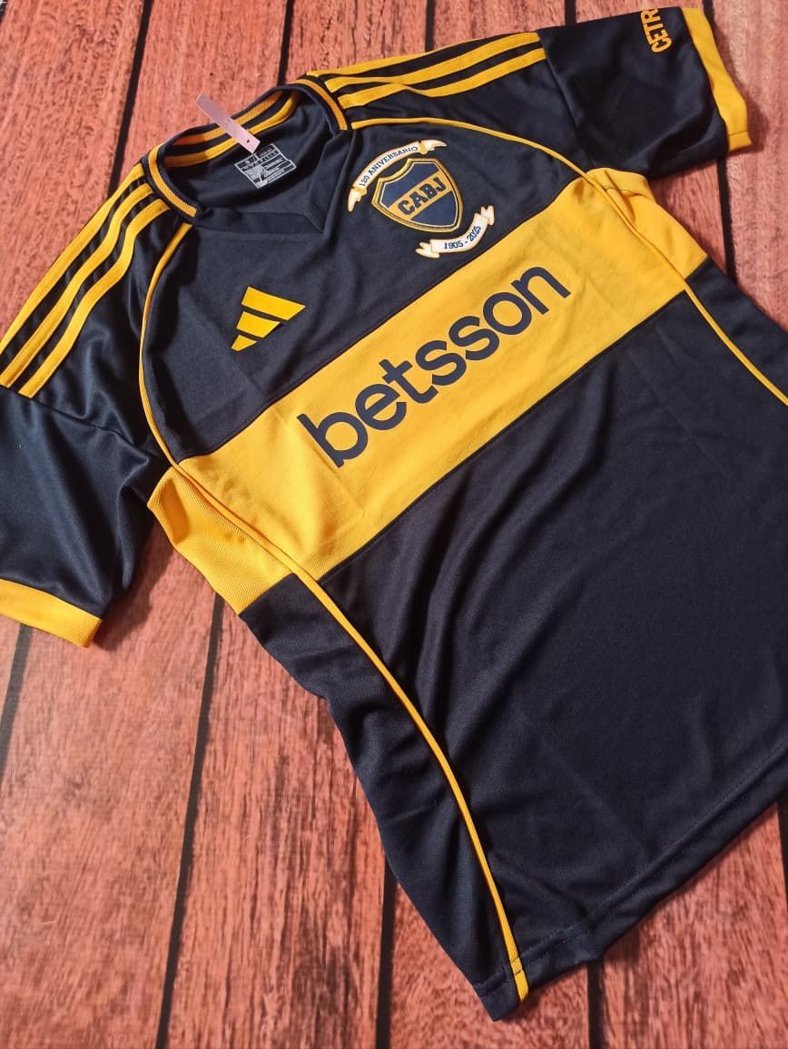 Una camiseta de fútbol negra y amarilla con el patrocinador Betsson, tendida sobre un suelo de madera.