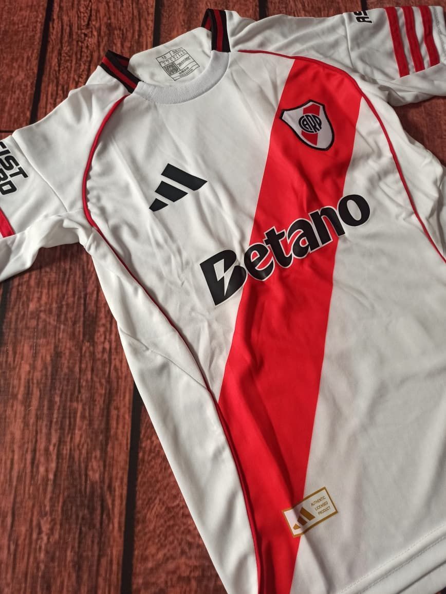Camiseta blanca y roja del River Plate con el patrocinador “Betano”, tendida sobre un suelo de madera.
