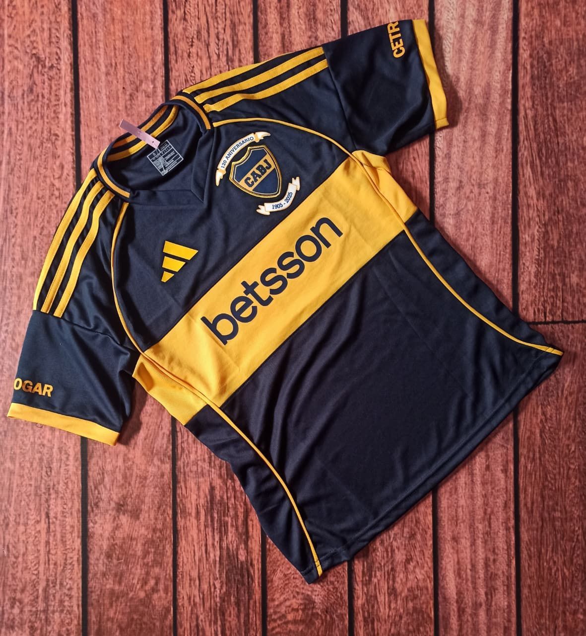 Una camiseta de fútbol negra y amarilla con el patrocinador Betsson, tendida sobre un suelo de madera.