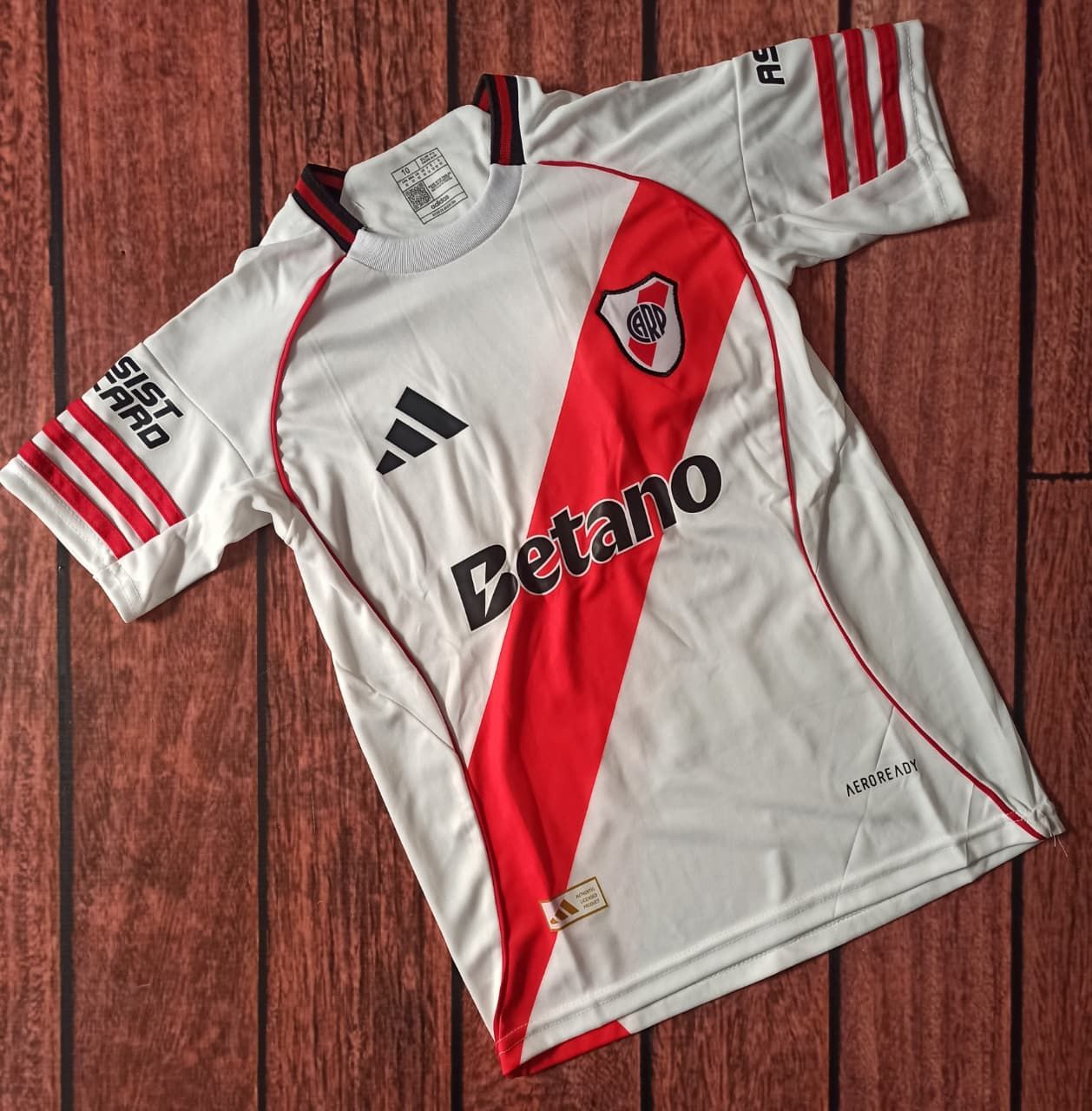 Camiseta blanca y roja del River Plate con el patrocinador “Betano”, tendida sobre un suelo de madera.