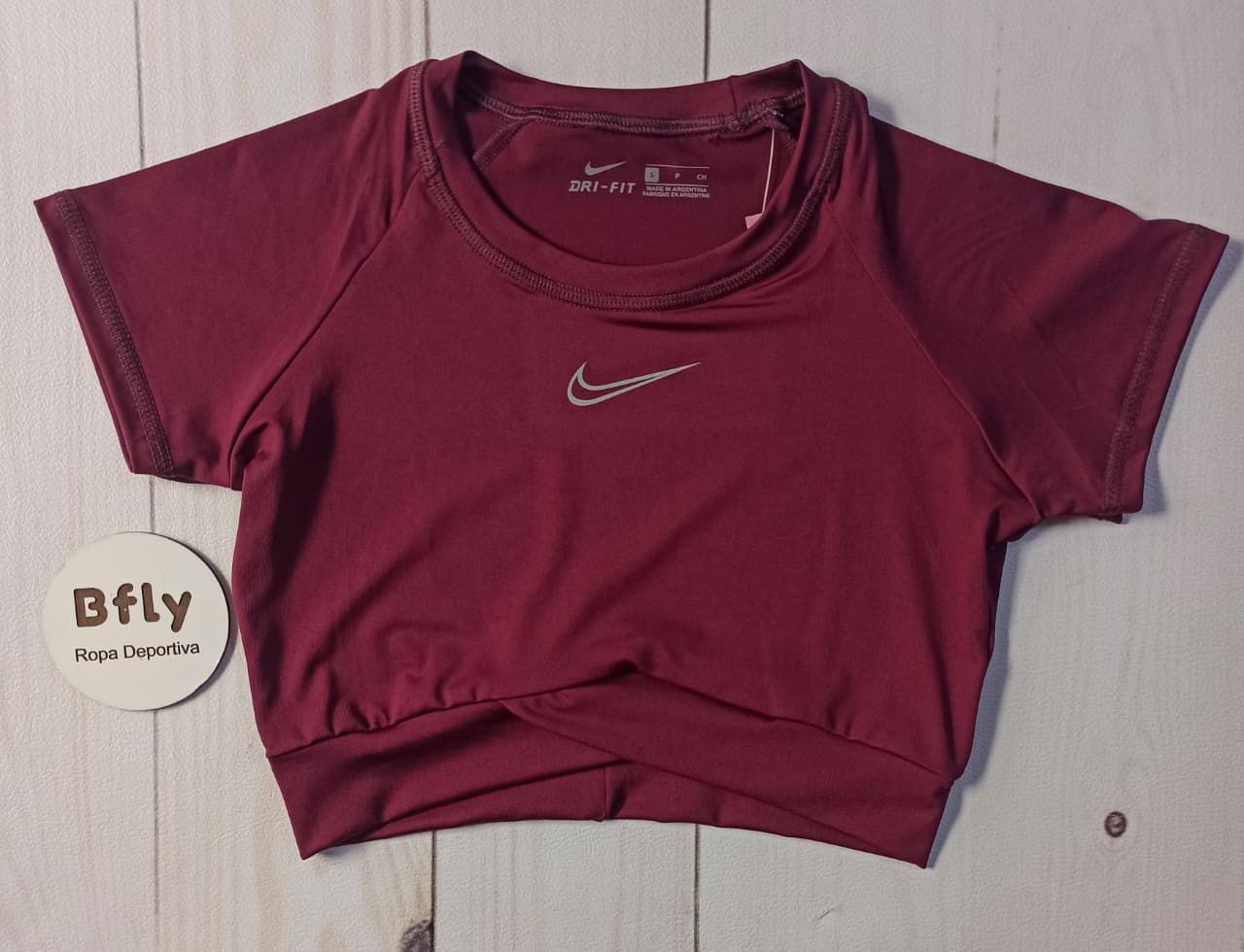 Tres tops cortos Nike doblados en rosa palo, marrón y magenta sobre una superficie de madera clara.