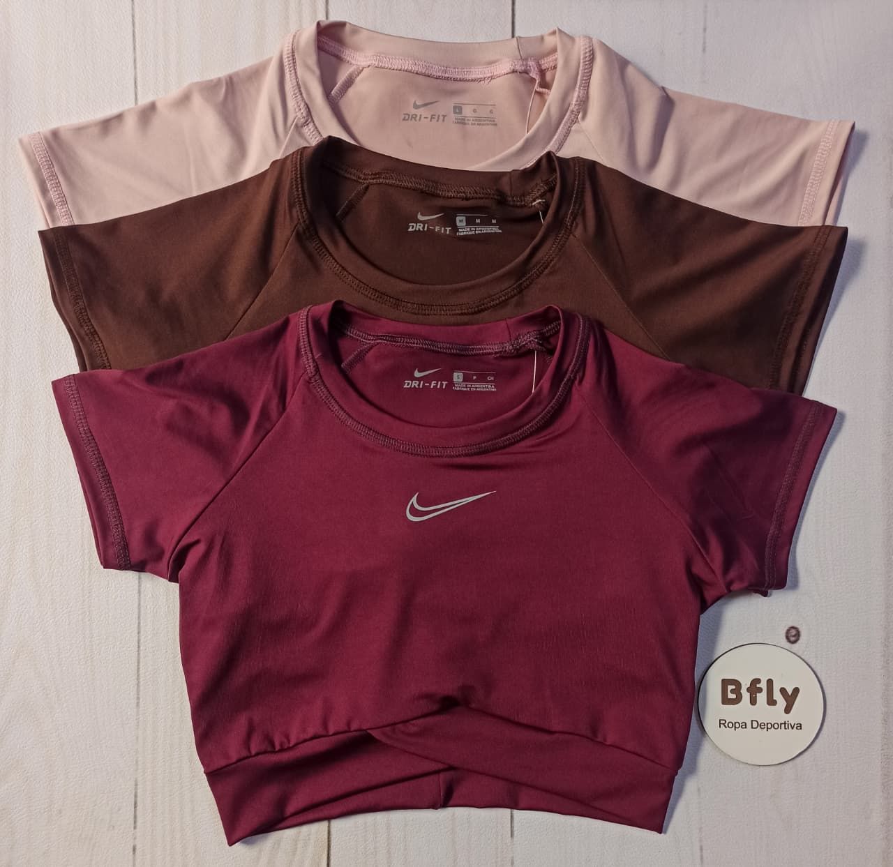 Tres tops cortos Nike doblados en rosa palo, marrón y magenta sobre una superficie de madera clara.