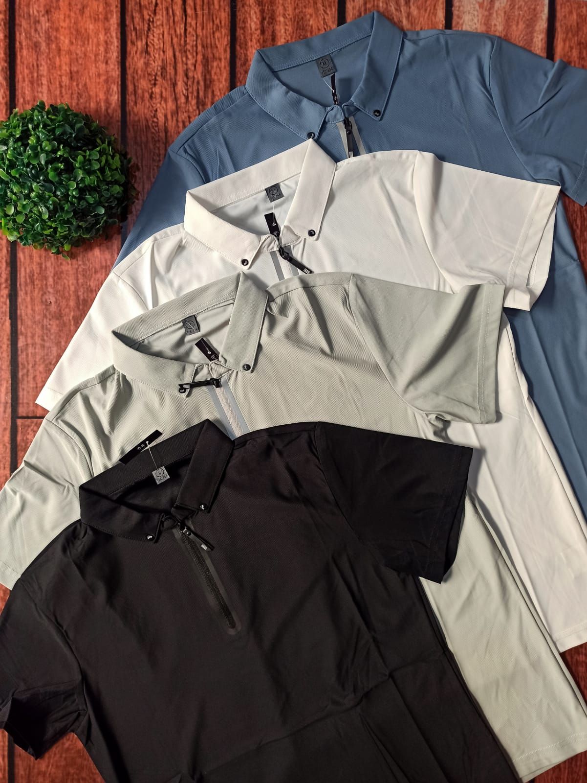 Cuatro camisas abotonadas dobladas en negro, beige, blanco y azul sobre un suelo de madera con una pequeña planta verde.